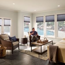 A man sitting in a sunroom with shade partially raised, revealing a view of the pool and backyard.