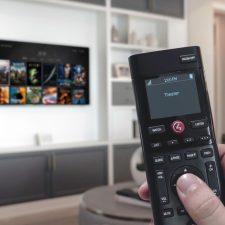 A close up of a Control4 remote control with a blurred out TV and media cabinets in the background.