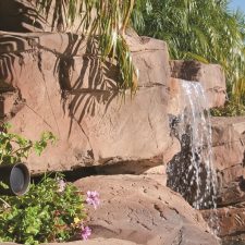 Landscape speaker installed among plants beside a backyard waterfall.